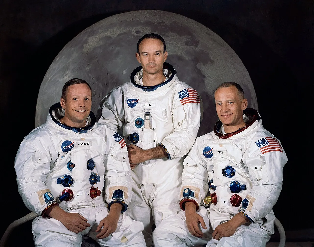 The Apollo 11 prime crew in white space suits before a lunar backdrop: Neil Armstrong, Michael Collins, and Buzz Aldrin.