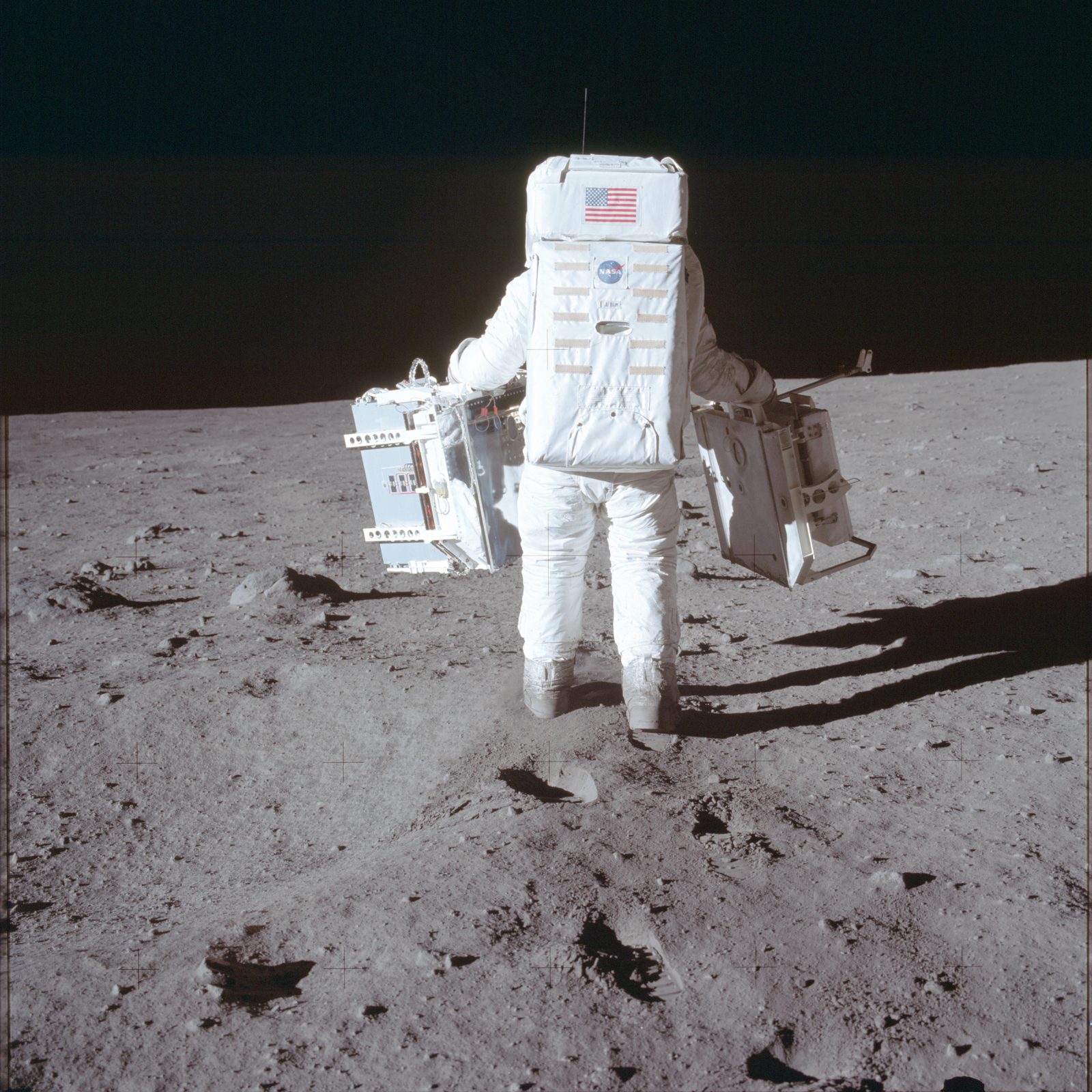 Aldrin carrying the two EASEP instruments — the Passive Seismic Experiment in his right hand, the Laser Ranging Retroreflector in his left — to the deployment site south of the LM (AS11-40-5942, NASA).